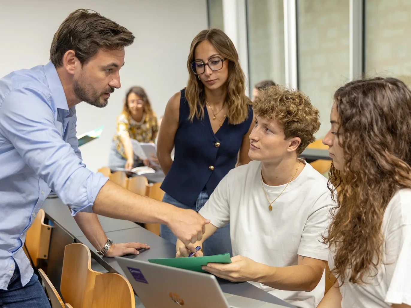 A teacher converses with three students seated in a modern classroom during a discussion about a computer assignment
